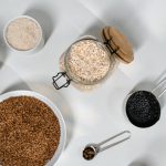Top view of various grains and seeds in bowls and jars on a white surface.