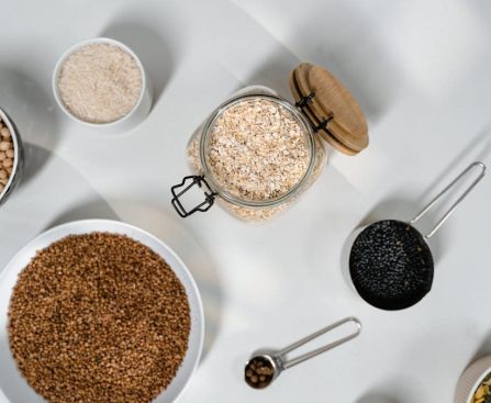 Top view of various grains and seeds in bowls and jars on a white surface.
